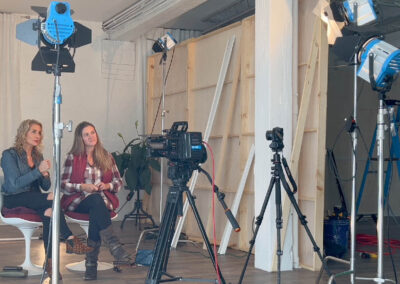 Behind the scenes of a video production on a set built at Flower City Studios. Lights and cameras in foreground and interviewees on a production set in the background