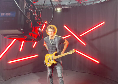 Man with guitar on a set with red lights floating in the background and camera in the foreground ready to film the guitarist playing